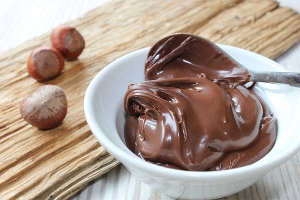 World Nutella Day. Chocolate spread in a bowl with a spoon on it. Bowl on a wooden surface with 3 hazelnuts beside it.