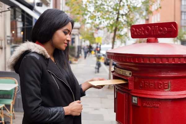 Send A Card To A Friend Day. Lady posting a card into a red Royal Mail post box