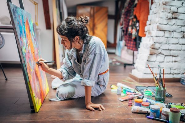 Girl sitting on the ground surrounded by colourful paint and painting on a canvas for International Artists Day