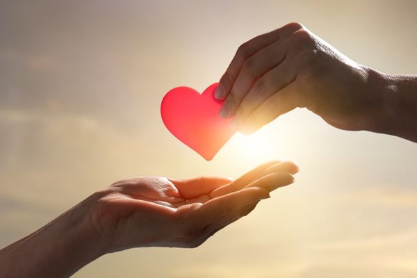 One hand passing a red heart to another hand against a sunset background for International Day of Charity