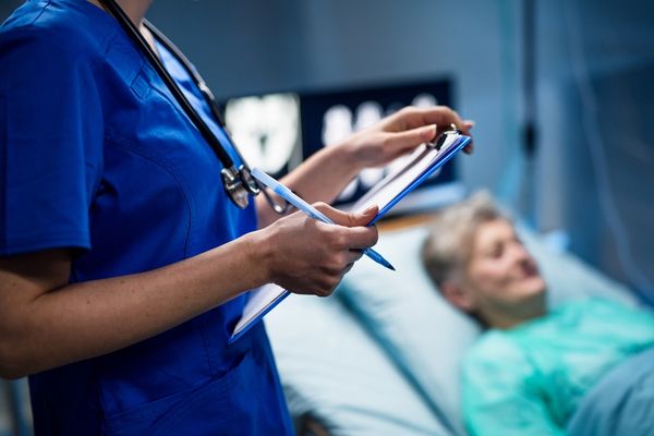 Nurse holding a clipboard with a patient lying in the background for International Infection Prevention Week