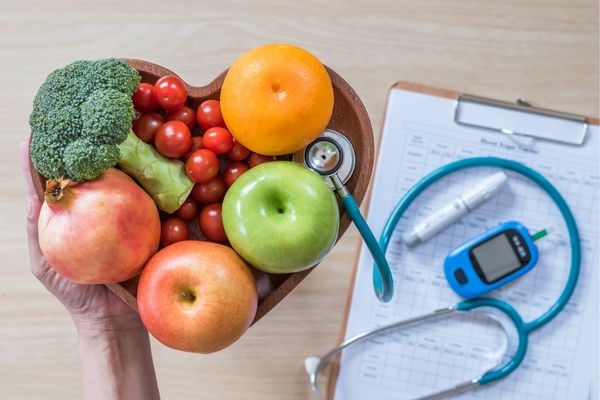 Hearts-shaped bowl of fruit with a clipboard and stethoscope for National Cholesterol Month