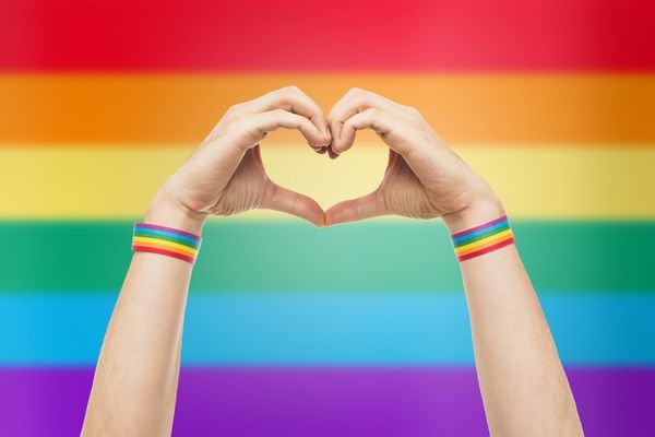 Two hands making a heart shape in front of a rainbow background for National Coming Out Day