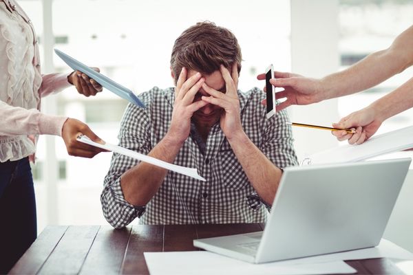 Man sitting at a desk with his head on his hands, people handing him phones and paperwork for National Stress Awareness Day