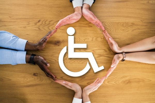Wooden blackground, 4 peoples hands linked together around a white symbol of a person in a wheelchair for International Day of Persons with Disabilities