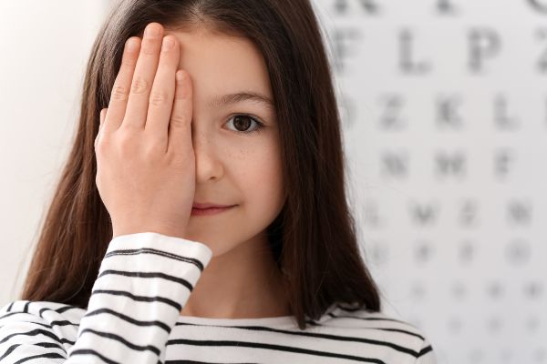 Child covering her eye standing in front of an eye chart in an opticians for Myopia Action Month